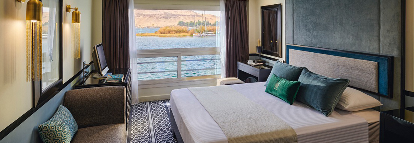 A refined guest cabin on the MS Hamees with a large window overlooking the Nile riverbank.