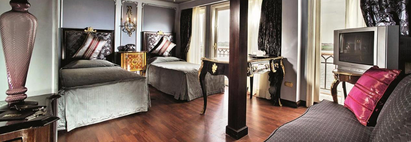 A luxury suite on the Mövenpick SS Misr with a large window, brass bedframe, and period-accurate furniture.